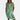 
A woman wearing a green, one-piece tropical print summer dress with floral patterns, featuring short sleeves and pockets. She is standing with one arm slightly raised, highlighting the fit around the waist and shoulder. The dress is designed to be comfortable for warm weather, showcasing a flowy style that reaches just above the knees.