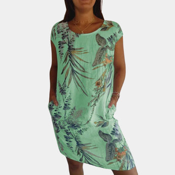 
A woman wearing a green, one-piece tropical print summer dress with floral patterns, featuring short sleeves and pockets. She is standing with one arm slightly raised, highlighting the fit around the waist and shoulder. The dress is designed to be comfortable for warm weather, showcasing a flowy style that reaches just above the knees.